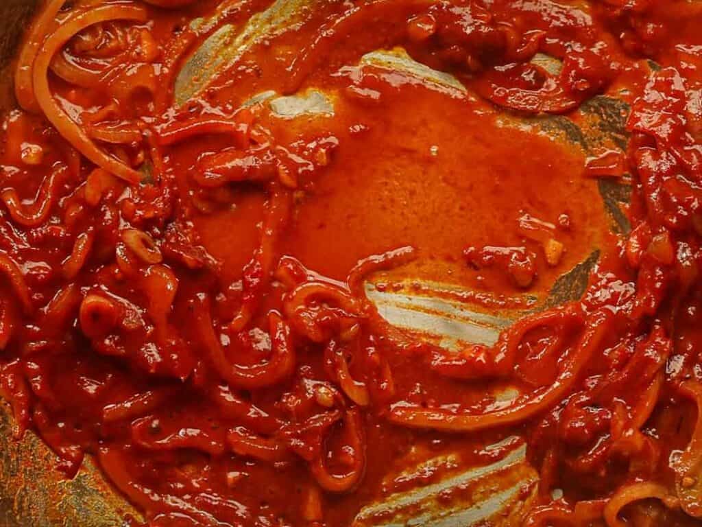 Close-up of a rich, red tomato sauce with shallots and garlic. The sauce appears to be simmering, with a glossy texture and visible streaks where its been stirred in a pan.